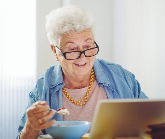 Elderly woman reading while eating a meal in her kitchen
