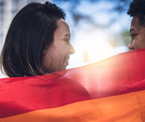 Two people smiling and holding up a rainbow Pride flag behind their shoulders.jpg