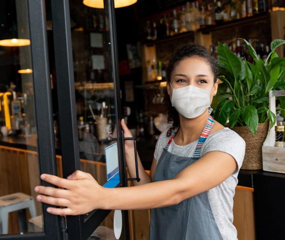 Woman business owner wearing a mask opening the door to her bar