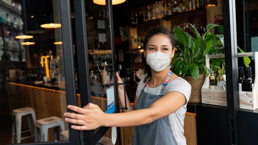 Woman business owner wearing a mask opening the door to her bar