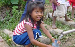 Flicka som sitter på huk och får rent vatten ur ett rör
