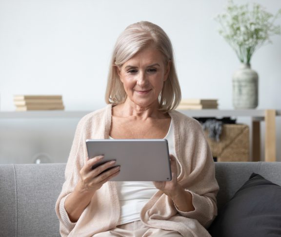 Portrait of pleasant middle aged woman sitting on cozy sofa, using tablet computer at home. Happy female retiree web surfing information, shopping in popular online store, reading electronic book.