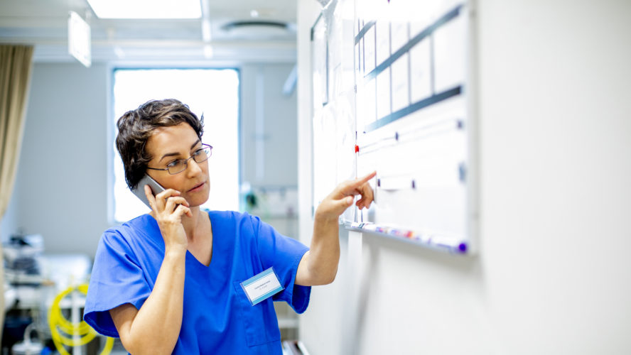 Young female doctor standing in the corridor of a modern hospital reading a scheduling whiteboard and talking on a cellphone