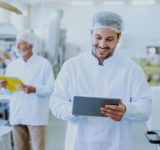 Young Caucasian smiling supervisor in sterile white uniform using tablet while standing in food plant