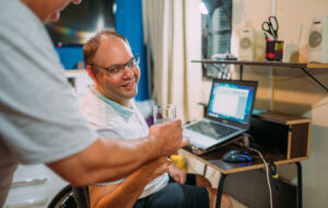 man in wheelchair using laptop at home. Father taking water for his son.