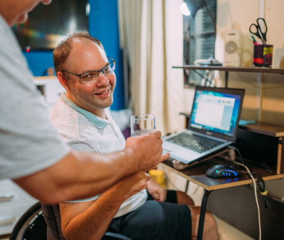 man in wheelchair using laptop at home. Father taking water for his son.