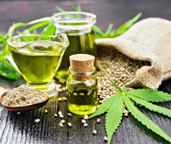 Hemp oil in two glass jars and sauceboat with grain in the bag, leaves and stalks of cannabis, a spoon with flour on the background of wooden boards
