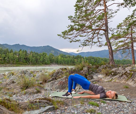 Blond beautiful girl is making a warm-up lying on the stone ground in the pose of the bridge, resting his palms and shoulder blades outdoors in the mountains near the river. Workout in nature.