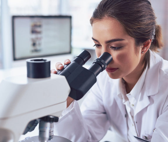 Shot of a cheerful young female scientist looking through the lens of a microscope while being seated inside of a laboratory