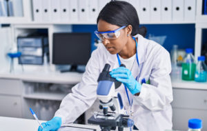 Young african american woman scientist using microscope write on document at laboratory