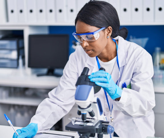 Young african american woman scientist using microscope write on document at laboratory