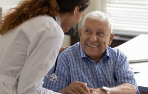 Friendly young female doctor and optimistic elder 80s man meeting in office for consultation. GP therapist holding hands of old patient, giving sympathy, support, advice about treatment