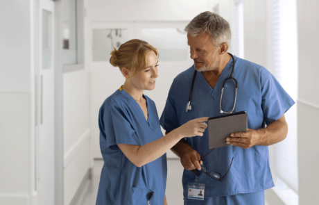 Medical Team Wearing Scrubs Meeting In Hospital Corridor Looking At Digital Tablet Together