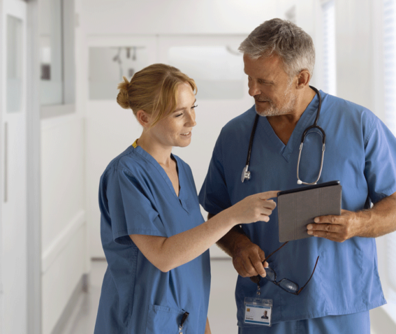 Medical Team Wearing Scrubs Meeting In Hospital Corridor Looking At Digital Tablet Together