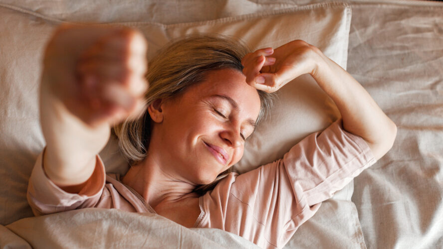 Good morning, new day, weekend, holiday. Happy middle aged woman lying on bed, lady stretching arms after sleep and enjoying morning in cozy comfort bedroom interior, free space