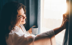 Curly haired lady in white bathrobe opens curtains to look outside large window drinking hot coffee in dark hotel room at sunrise time