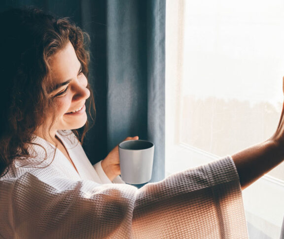 Curly haired lady in white bathrobe opens curtains to look outside large window drinking hot coffee in dark hotel room at sunrise time