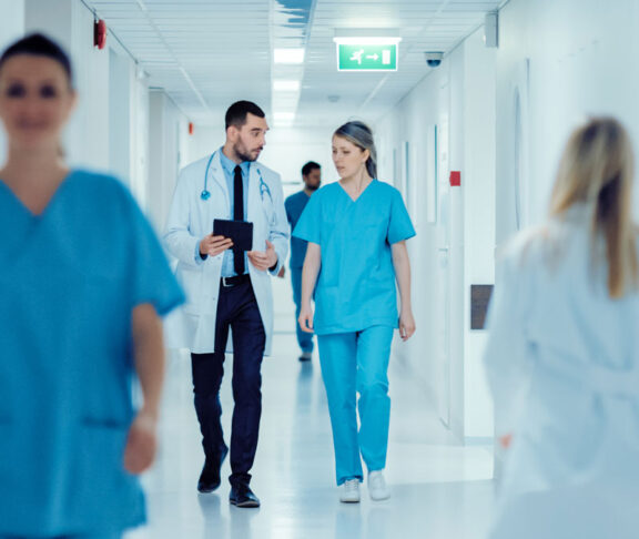 Surgeon and Female Doctor Walk Through Hospital Hallway, They Consult Digital Tablet Computer while Talking about Patient's Health. Modern Bright Hospital with Professional Staff