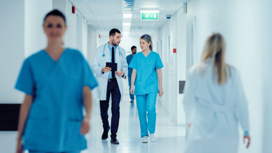 Surgeon and Female Doctor Walk Through Hospital Hallway, They Consult Digital Tablet Computer while Talking about Patient's Health. Modern Bright Hospital with Professional Staff