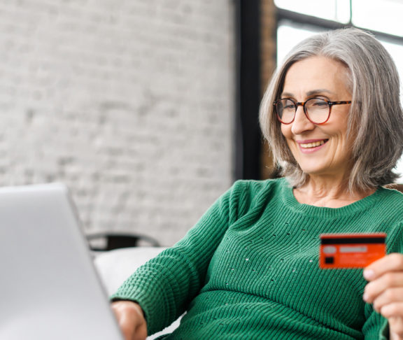 Pretty senior businesswoman in glasses and green jumper sitting at the desk using laptop and credit card for online payment from home, typing number to enter internet banking account, ordering food