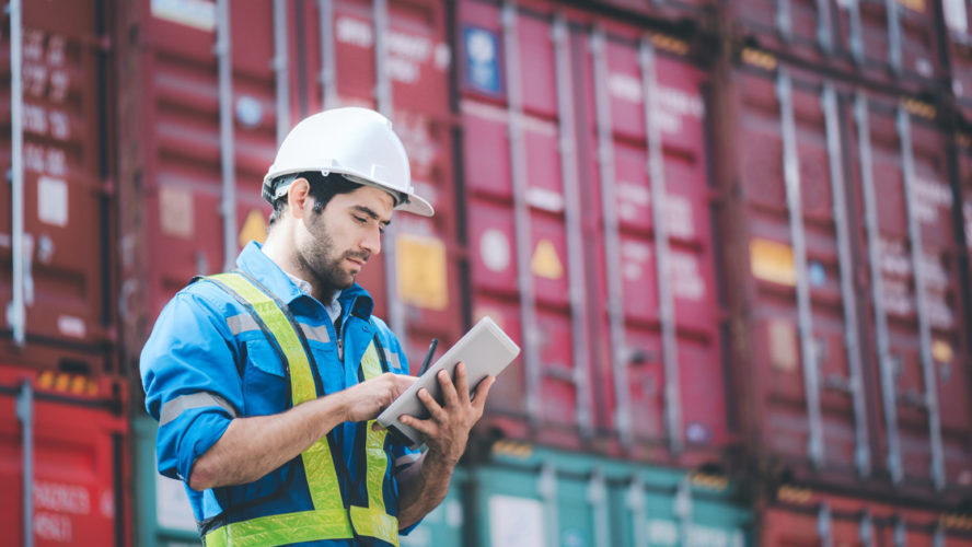 Man wears hardhat and reflection shirt and checking tablet with blurry metal containers in background. Concept of inventory and logistic management.