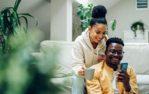 Multiracial young couple sitting on a comfortable sofa using smartphone and holding cups of hot beverages. Spouse relaxing together on cozy couch and online shopping on a mobile phone. Copy space.