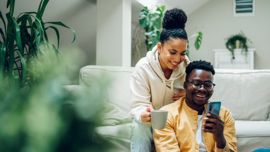 Multiracial young couple sitting on a comfortable sofa using smartphone and holding cups of hot beverages. Spouse relaxing together on cozy couch and online shopping on a mobile phone. Copy space.