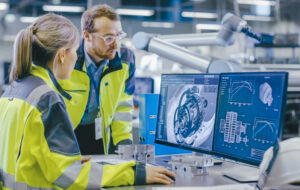 At the Factory: Male Mechanical Engineer Holds Component while Female Chief Engineer Work on Personal Computer, They Discuss Details of the 3D Engine Model Design. - stock photo