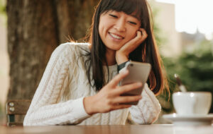 Happy young woman sitting at outdoor cafe and using her cellphone. Female model reading text message on smart phone at coffee shop and smiling.