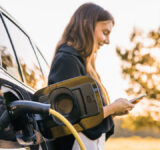 Girl charging electric car parked in the nature area and adjusting an EV charging app on smartphone