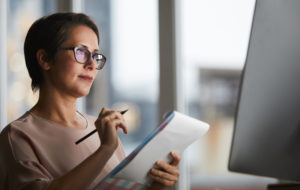 Businesswoman with document looking at computer screen and making working notes in office