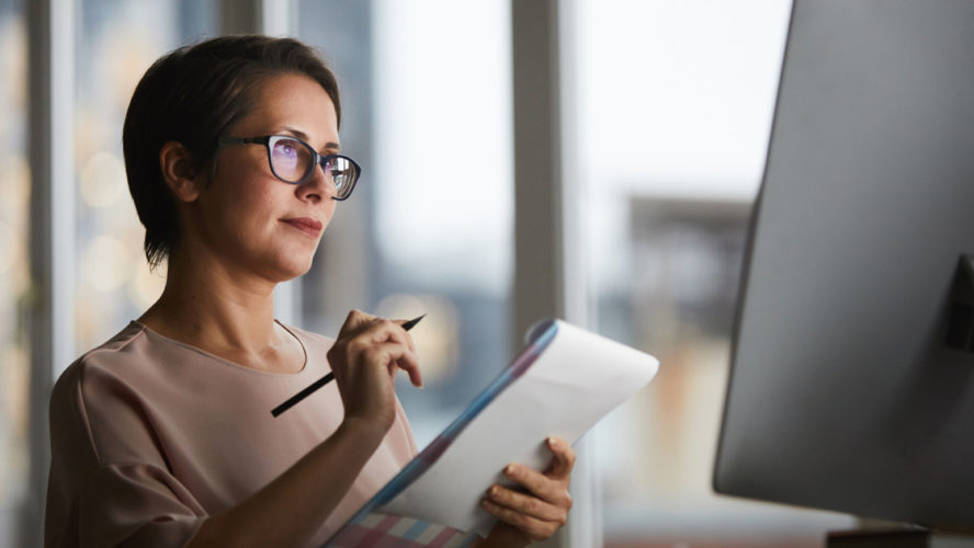 Businesswoman with document looking at computer screen and making working notes in office
