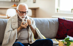 Senior man suffering from flu drinking tea while sitting wrapped in a blanket on the sofa at home.
