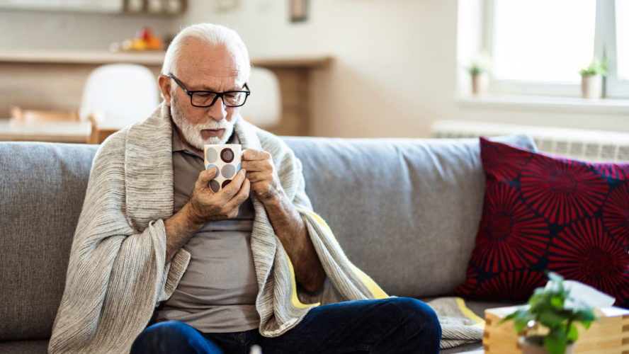 Senior man suffering from flu drinking tea while sitting wrapped in a blanket on the sofa at home.