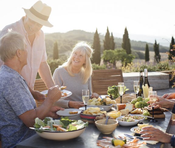 A group of mature friends are sitting around an outdoor dining table, eating and drinking. They are celebrating their holiday with a glass of champagne and enjoying each others company. The image has been taken in Tuscany, Italy.