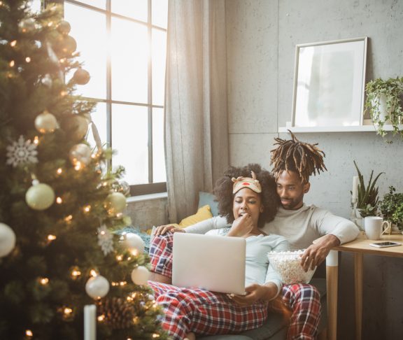 Young couple celebrating Christmas at home. Home is decorated with Christmas ornaments and lights, they sitting on sofa and using laptop