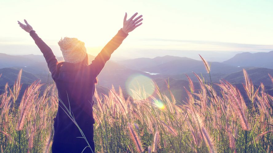 Kvinde står på toppen af bjerg og strækker armene imod solen