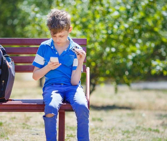 Dreng sidder på en bænk med sin rygsæk og kigger på sin telefon