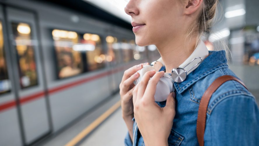 Unrecognizable young woman in denim shirt with earphones, standing at the underground platform, waiting to enter a train