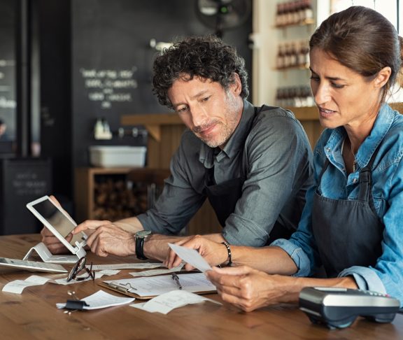 Man and woman sitting in cafeteria discussing finance for the month. Stressed couple looking at bills sitting in restaurant wearing uniform apron. Café staff sitting together looking at expenses and bills.