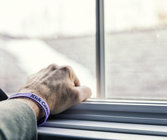 A senior adult man cancer patient - about to start a three month chemotherapy regimen - is clenching his fist with his hand resting on a window sill - looking out toward the blurry and indistinct future outside the window. He is wearing a generic cancer themed wristband (personally "designed" by me - the photographer/patient) with the embossed words "Kick Cancer".
