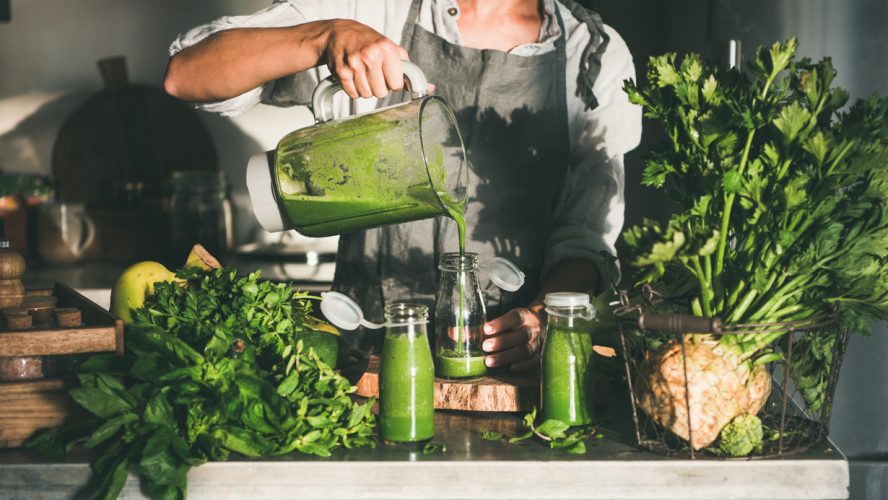 Making green detox take-away smoothie. Woman in linen apron pouring green smoothie drink from blender to bottle surrounded with vegetables and greens. Healthy, weight loss food concept