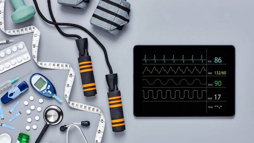 Directly above flat lay of medical equipment and digital tablet. Glucometer jump rope bandages and tape measure are diagonally placed with medicines. Computer screen is displaying pulse trace.