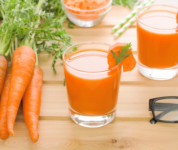 Two glasses of fresh carrot juice, bunch of carrot with green and black-rimmed spectacles on light wooden background. Concept carrot for good eyesight. Horizontal orientation.