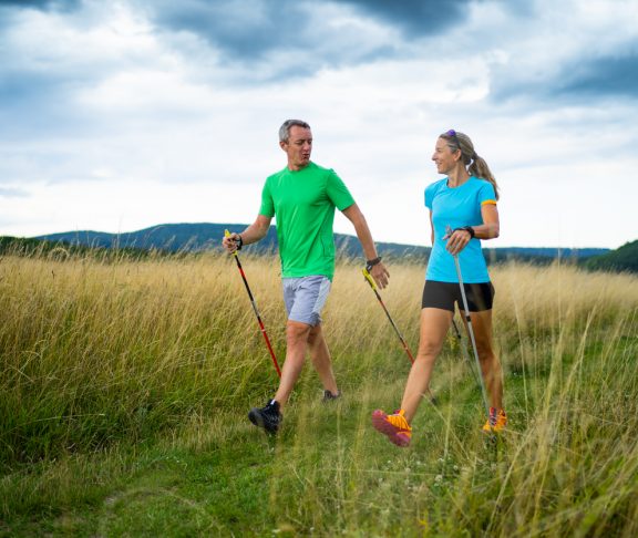 active smiling middle aged couple doing nordic walking sport in grassland with shallow focus cloudy overcast sky dark clouds front view