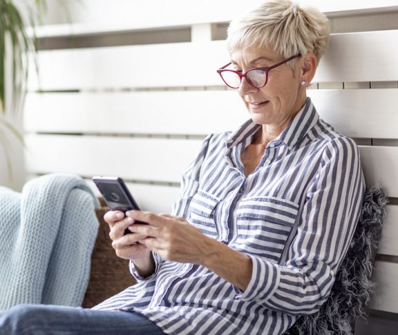 Mature woman with mobile phone on her hands sitting in room and sending messages to her friends and family