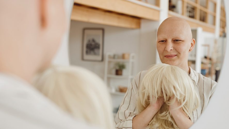 Syöpää sairastava nainen katsoo peiliin ja pitelee aidon näköistä peruukkia