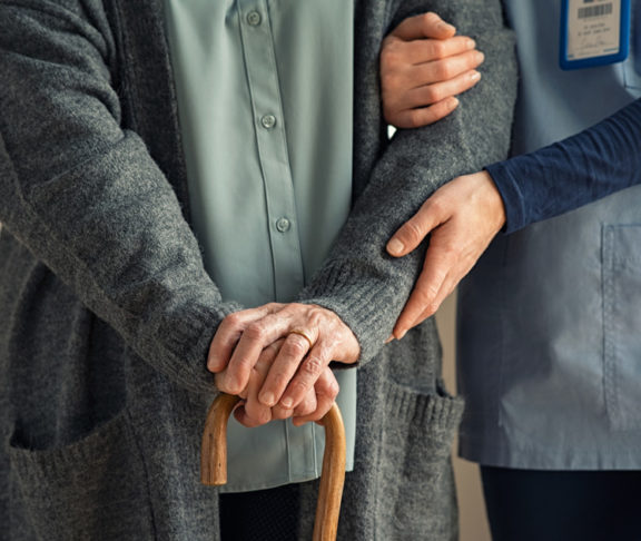 Close up hands of caregiver doctor helping old woman at private clinic. Close up of hands of nurse holding a senior patient with walking stick. Elder woman using walking cane at nursing home with nurse holding hand for support.