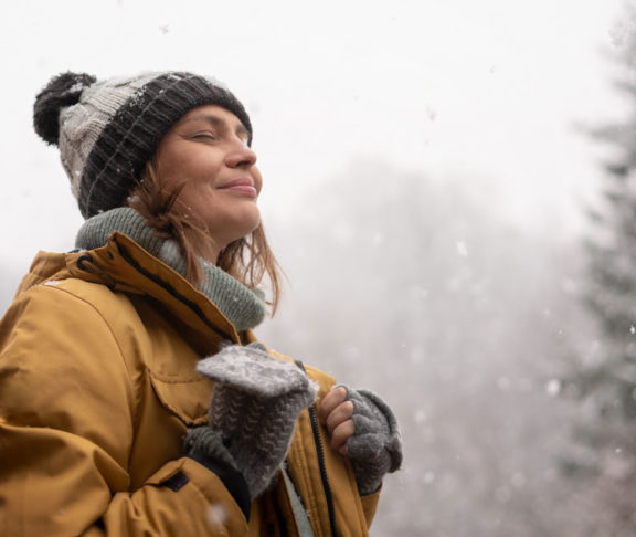 Young mature adult caucasian woman in a hat and yellow jacket breathing fresh air in the winter forest