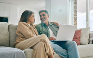 Happy middle aged couple using laptop computer relaxing on couch at home. Smiling mature man and woman talking having fun laughing with device sitting on sofa in sunny living room. Candid shot